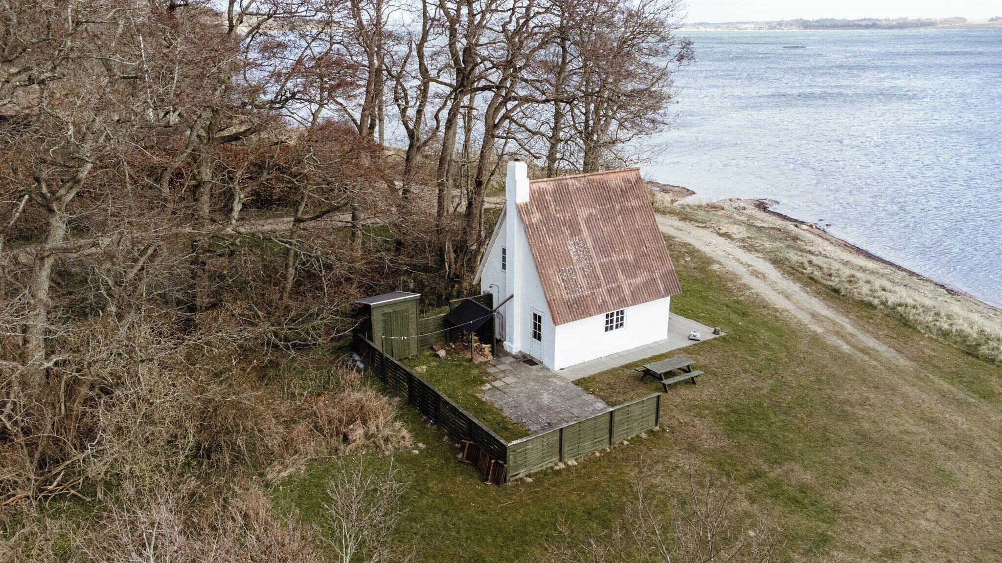 En hvid bygning med skråt tag beliggende ved vandet, omgivet af træer og græs.
