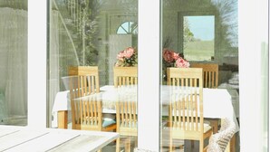 Uma área de refeições com cadeiras de madeira e uma mesa, uma janela com vista para vegetação, e uma porta com um vitral circular.