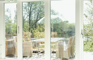 Habitació amb grans finestres, mobiliari de vimet i vista a un jardí i arbres.