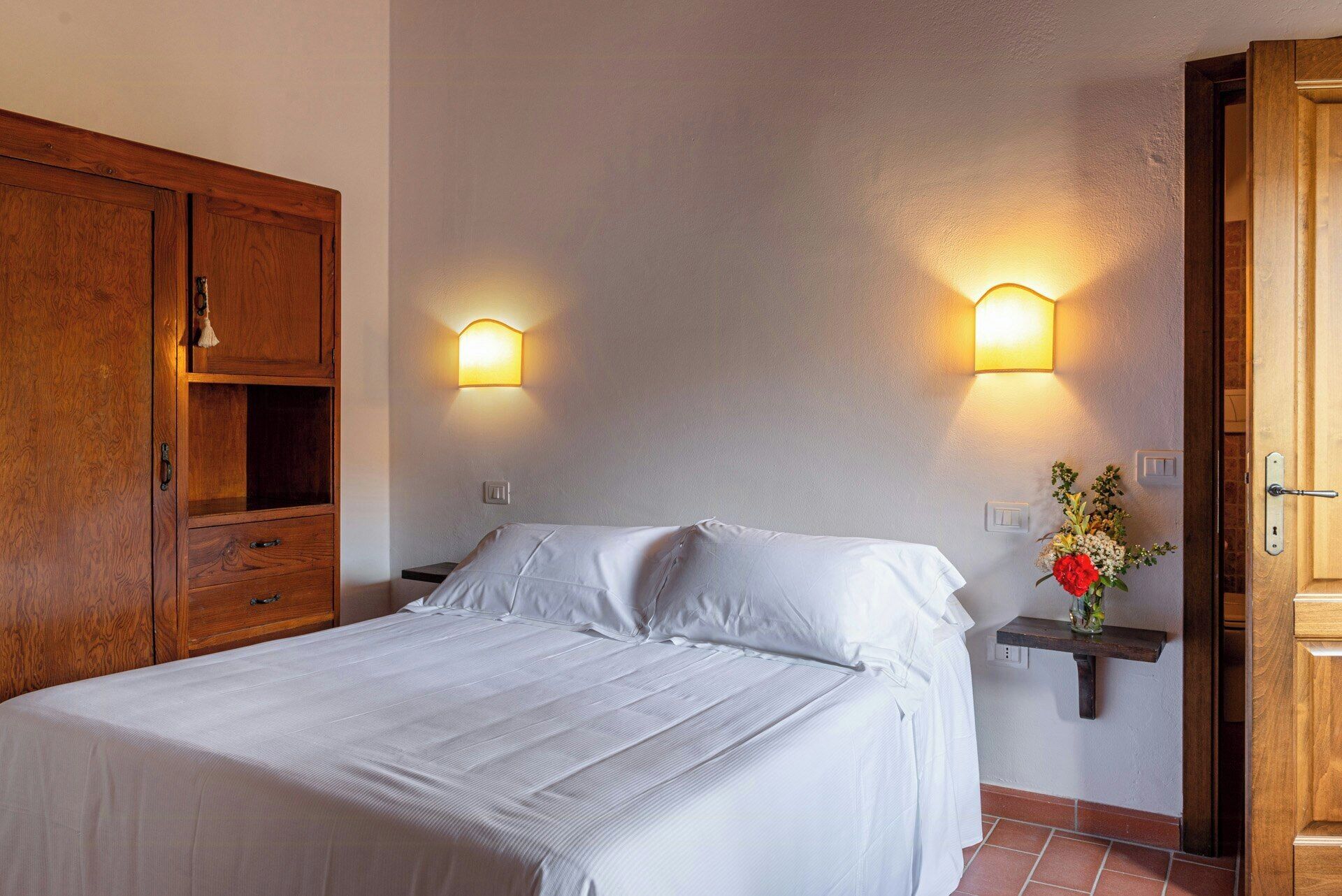 Ein Schlafzimmer mit einem Bett, einem Holzschrank, zwei Wandlampen, einem Nachttisch und einem kleinen Regal mit Blumen.