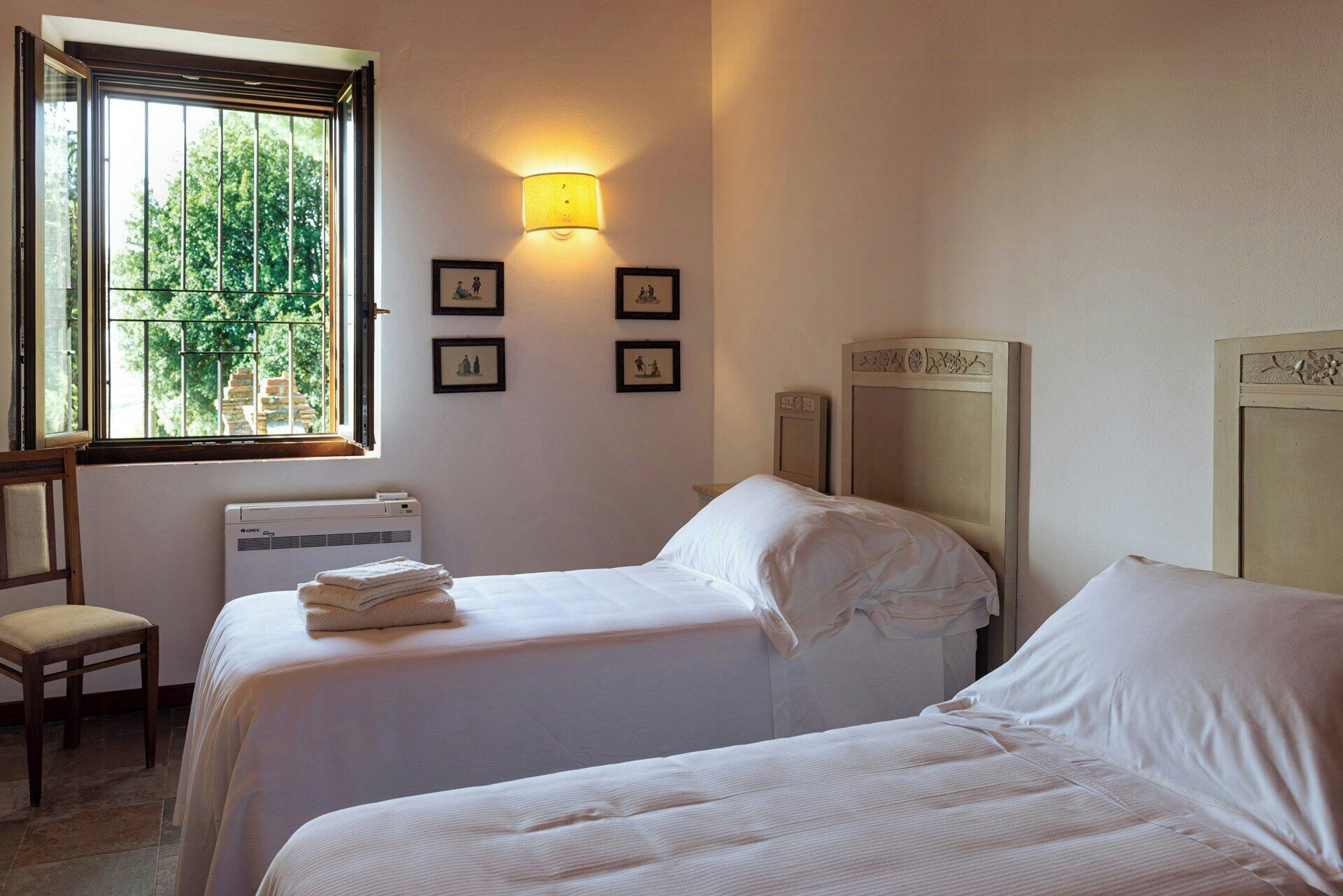 A hotel room with two beds, a window with a view of trees, a wall-mounted heater, and framed pictures.
