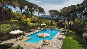 Una piscina rettangolare con piastrelle in pietra, circondata da lettini e ombrelloni, immersa in un rigoglioso giardino con alberi e vista su dolci colline.
