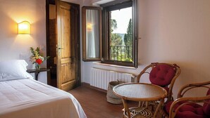 Camera d'albergo con un letto, un tavolino, sedie in vimini, una finestra con vista e una lampada a parete.