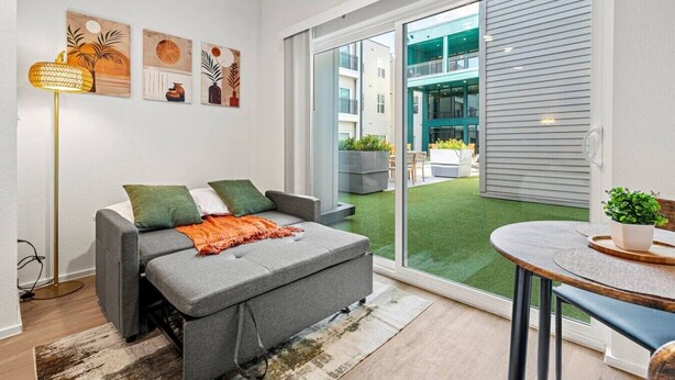 A modern living room with a grey sofa, a small round table, and a view of a green outdoor area through large sliding glass doors. - Bishop Arts Getaway | Yoga, Gym & Entertainment (Dallas)