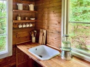 A wooden cabin kitchen with a window, wooden shelves, a sink, and a metal pitcher. - Off-grid cabin in your private Swedish forest (Simlångsdalen)