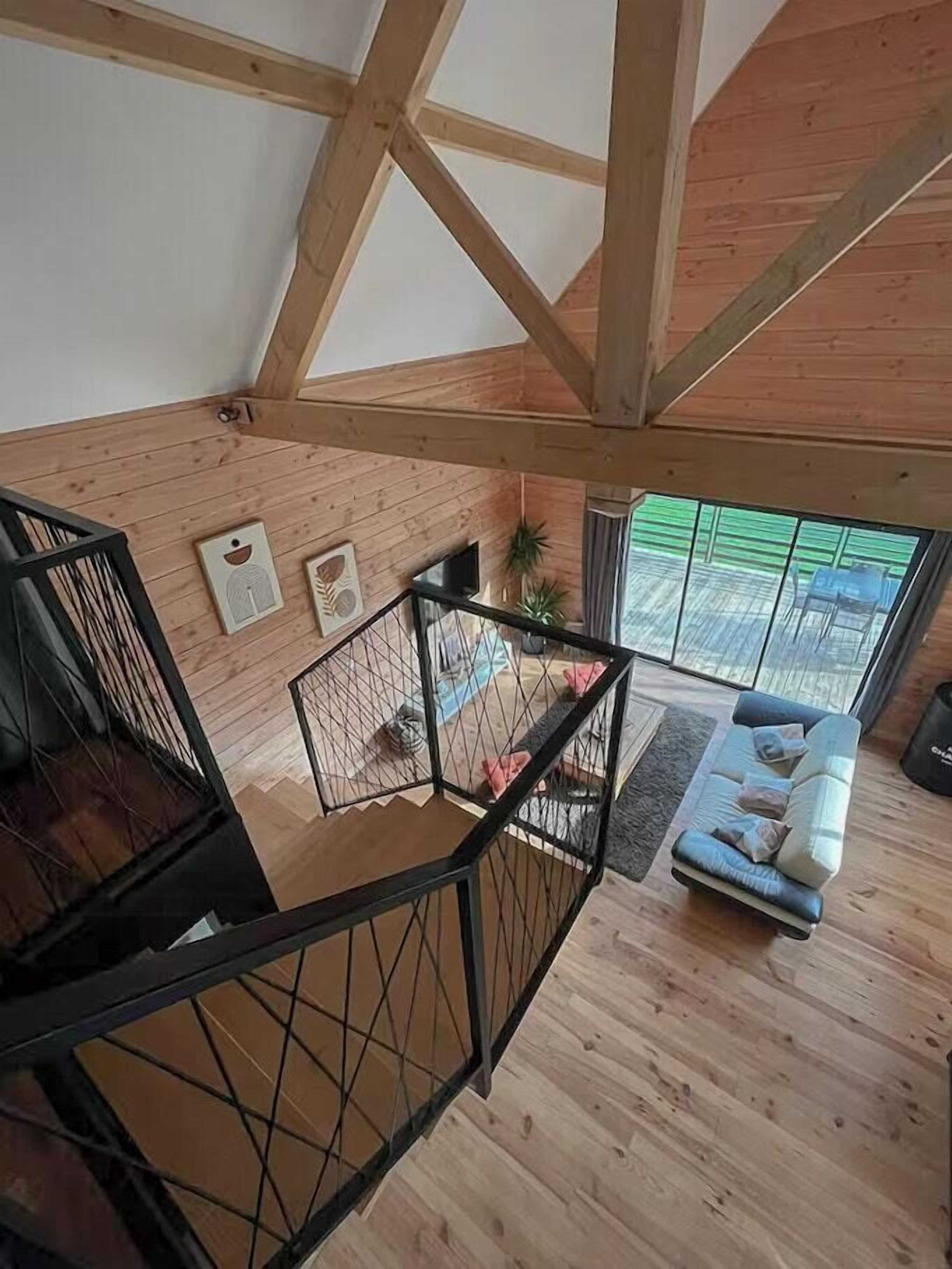Un dormitorio estilo loft con vigas de madera a la vista, una escalera de metal negro, un sofá azul y un ventanal con vistas a un espacio exterior verde.