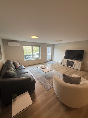 Ein modernes Wohnzimmer mit einer Couch, einem Sessel, einem Fernseher und Blick auf einen Balkon.