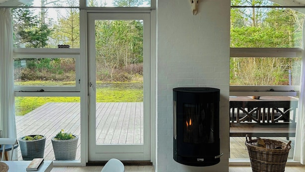 A room with a fireplace, a chair, a table, and a view through large windows. - Holiday home by Ålbæk Beach for the whole family (Ålbæk)