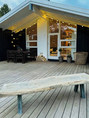 Eine Holzterrasse mit einer Bank, Stühlen und einem Tisch. Lichterketten und Kübelpflanzen sind vorhanden.