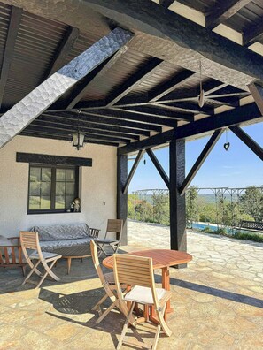 Eine überdachte Terrasse mit Tisch und Stühlen, einem Fenster und Blick auf Pool und Grünfläche.