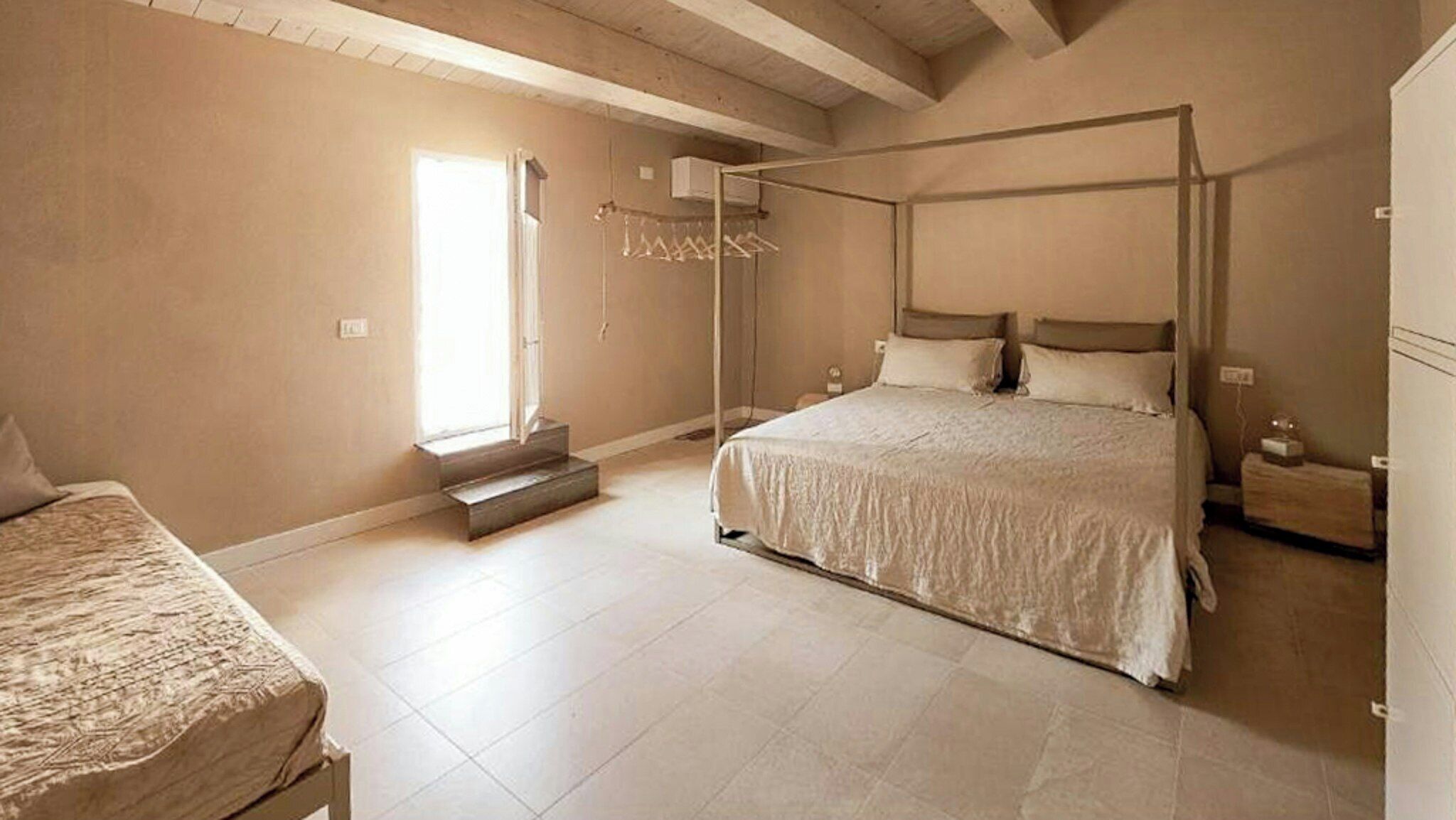 A bedroom with a four-poster bed, a bedside table, a nightstand, a window with curtains, and a built-in air conditioning unit.