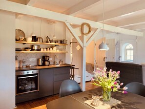 Eine moderne Küche mit Holzböden, einem Esstisch mit Blumenvase und einem schwarzen Sofa in einem hellen Raum mit weißen Wänden und Holzbalken.