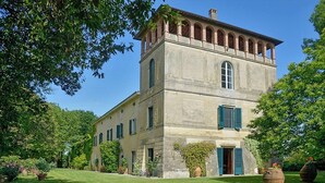 Un grand bâtiment en pierre à deux étages, doté d’une tour centrale, entouré d’une végétation luxuriante et de plantes en pot.