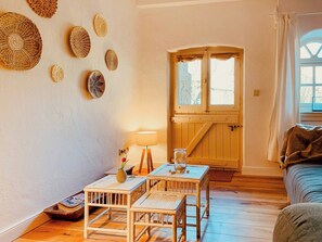 Ein gemütliches Zimmer mit Holzboden, einem kleinen Tisch mit Stühlen, einer Lampe und einem Fenster mit Vorhängen.