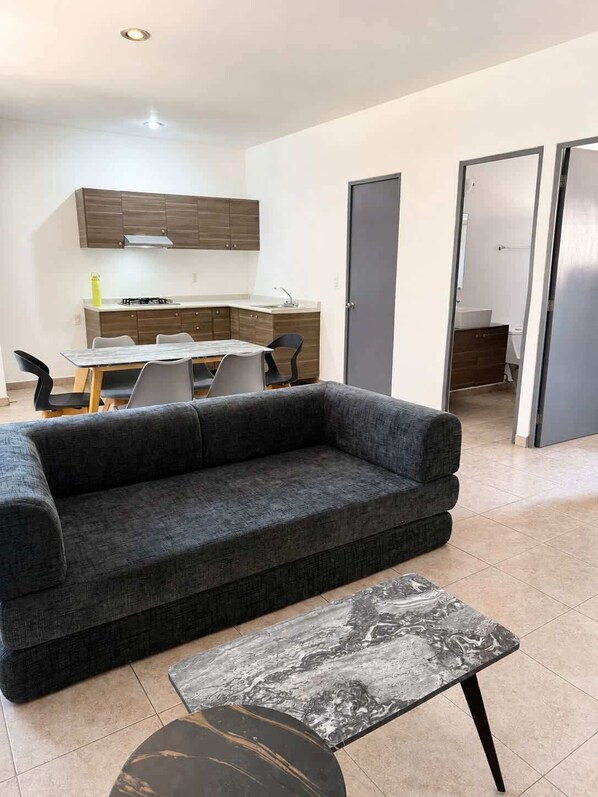 Een woonkamer met een grijze bank, een marmeren salontafel en een eethoek, met een keuken op de achtergrond.