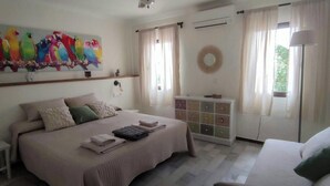 A bedroom with a bed, a dresser, a nightstand, a painting of colorful birds, and a window with curtains. - Ibizan design villa in Costa Blanca with pool (Torrevieja)