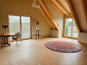 Ein geräumiges Zimmer mit Holzboden, einem Esstisch mit Stühlen, einer Vase mit Trockenblumen, einem roten Teppich mit Muster und großen Fenstern mit Blick auf Bäume.