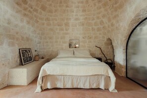 Ein Schlafzimmer mit Steinwand, einem Bett mit weißer Bettdecke, einem Nachttisch mit Lampe und einem Fenster mit Fliegengitter.
