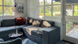 A modern living room with a gray sectional sofa, a small round table with a vase, and a door leading to a wooden deck. - Relax in the countryside with pool (Aulum)