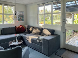 A modern living room with a gray sectional sofa, a small round table with a vase, and a door leading to a wooden deck. - Relax in the countryside with pool (Aulum)