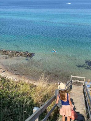 A person on a wooden staircase overlooking a beach with clear water and a distant boat. - The small, red wooden house with lots of charm (Vejby Strand)