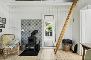 A cozy room with a wooden floor, a wicker chair, a black rug, a patterned wall, a stove, a ladder, and a door leading outside. - The small, red wooden house with lots of charm (Vejby Strand)