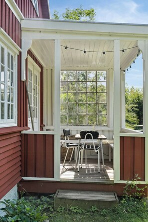 A covered porch with a table and chairs, a window with a view of greenery, and a string of lights. - The small, red wooden house with lots of charm (Vejby Strand)