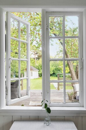 A window with a view of a garden and a white building. - The small, red wooden house with lots of charm (Vejby Strand)