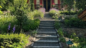 Ein Gebäude mit roter Tür, umgeben von einem Garten mit verschiedenen Pflanzen und einem Kiesweg, der zum Eingang führt.