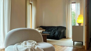 Ein Zimmer mit Holzboden, einer Couch, einem Sessel, einer Lampe und Blick durch ein Fenster.