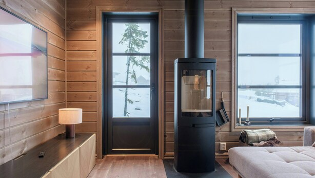 Una habitación con paredes de madera, una estufa de leña negra, una cama, una ventana con vistas nevadas y una puerta.