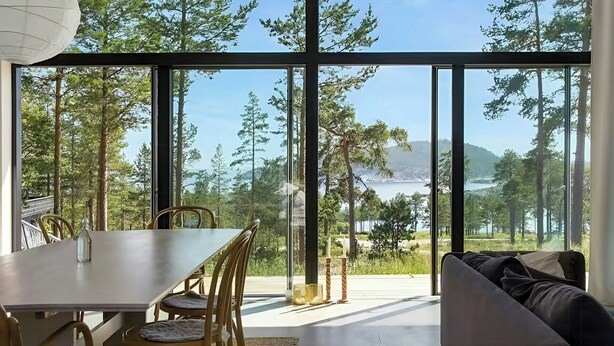 A modern interior with a dining area, a sofa, and large windows offering a view of a forest and a lake.