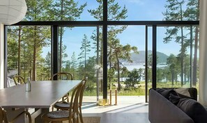 A modern interior with a dining area, a sofa, and large windows offering a view of a forest and a lake.