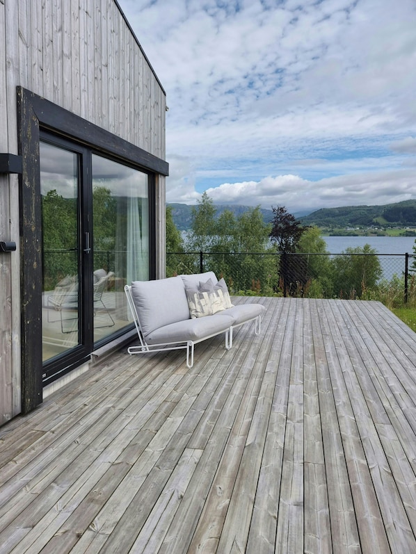 A modern deck with a sofa and glass doors overlooking a lake and hills.