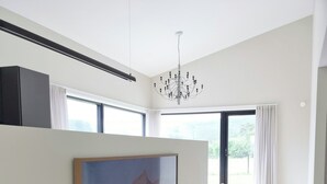 A modern living room with a large flat-screen TV mounted on the wall, a wooden entertainment unit below, a chandelier, and a view of the outdoors through large windows. - Cabin by the fjord with rooftop terrace on Randøy (Hjelmeland)