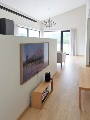 A modern living room with a large flat-screen TV mounted on the wall, a wooden entertainment unit below, a chandelier, and a view of the outdoors through large windows.