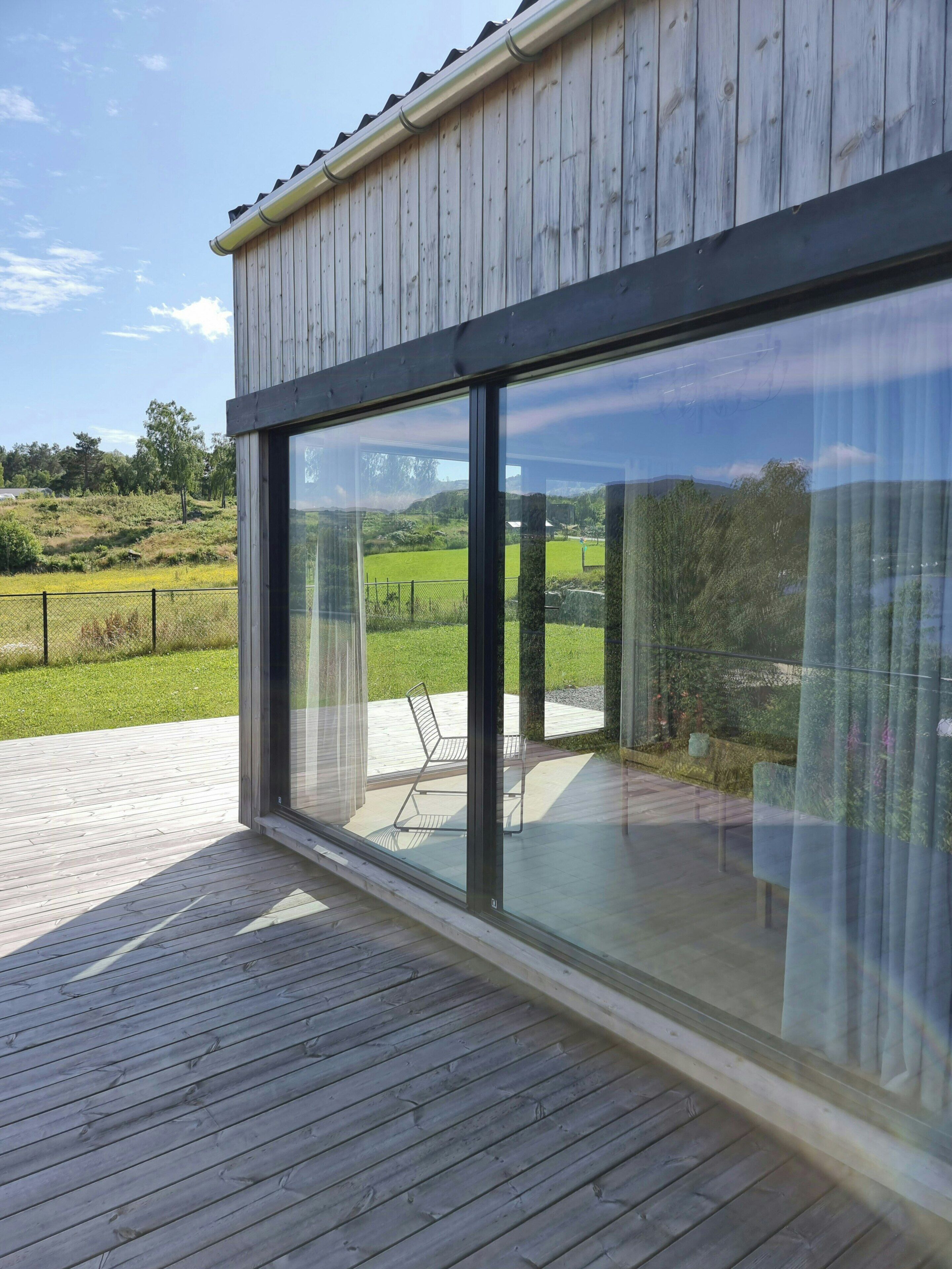 A modern building with large glass windows, a wooden deck, and a view of a green landscape.