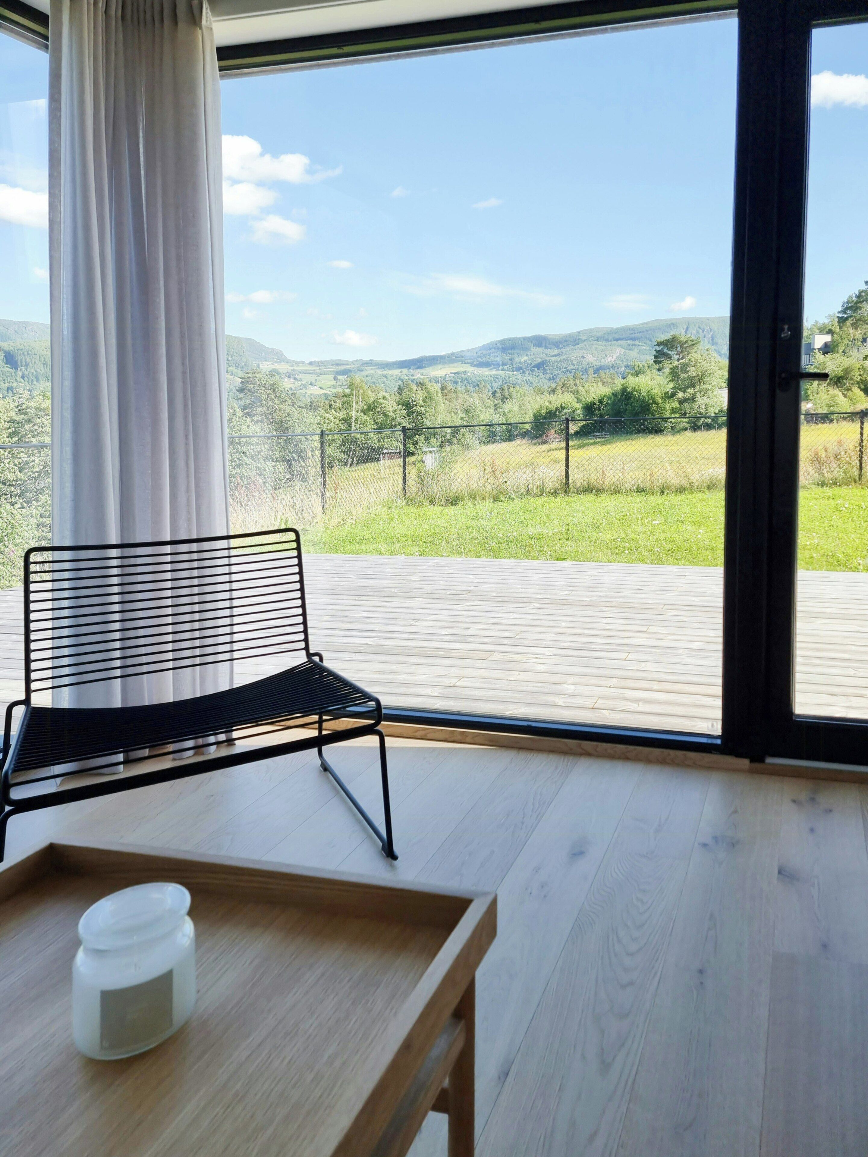A room with a view of a green landscape, a black metal chair, a wooden table, and a white container.