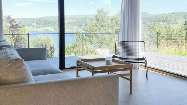 A modern living room with a sofa, a table, and a chair, large windows with a view of a lake and hills. - Cabin by the fjord with rooftop terrace on Randøy (Hjelmeland)