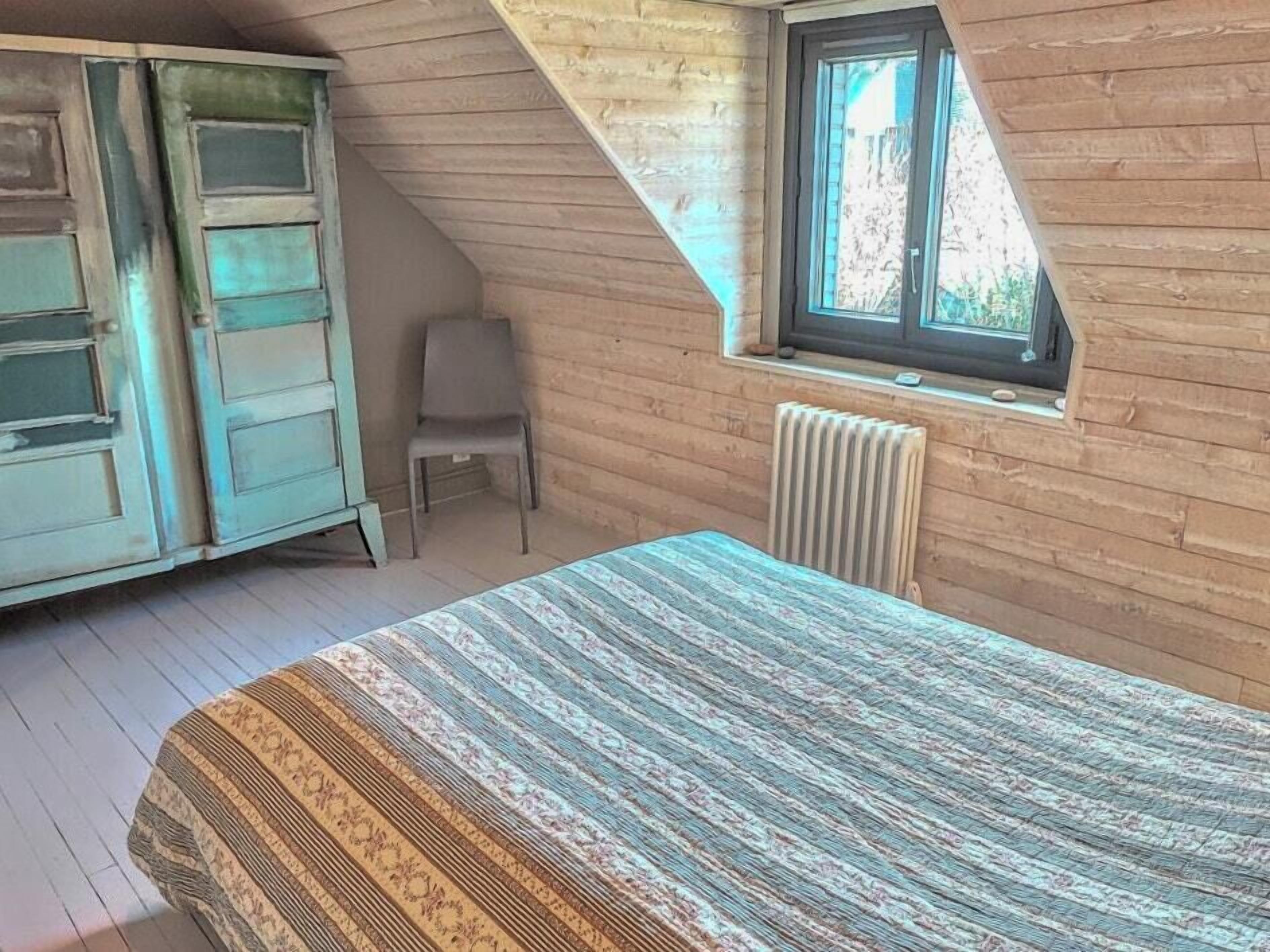 A room with a sloped ceiling, a window, a radiator, a bed with a patterned bedspread, and a chair.