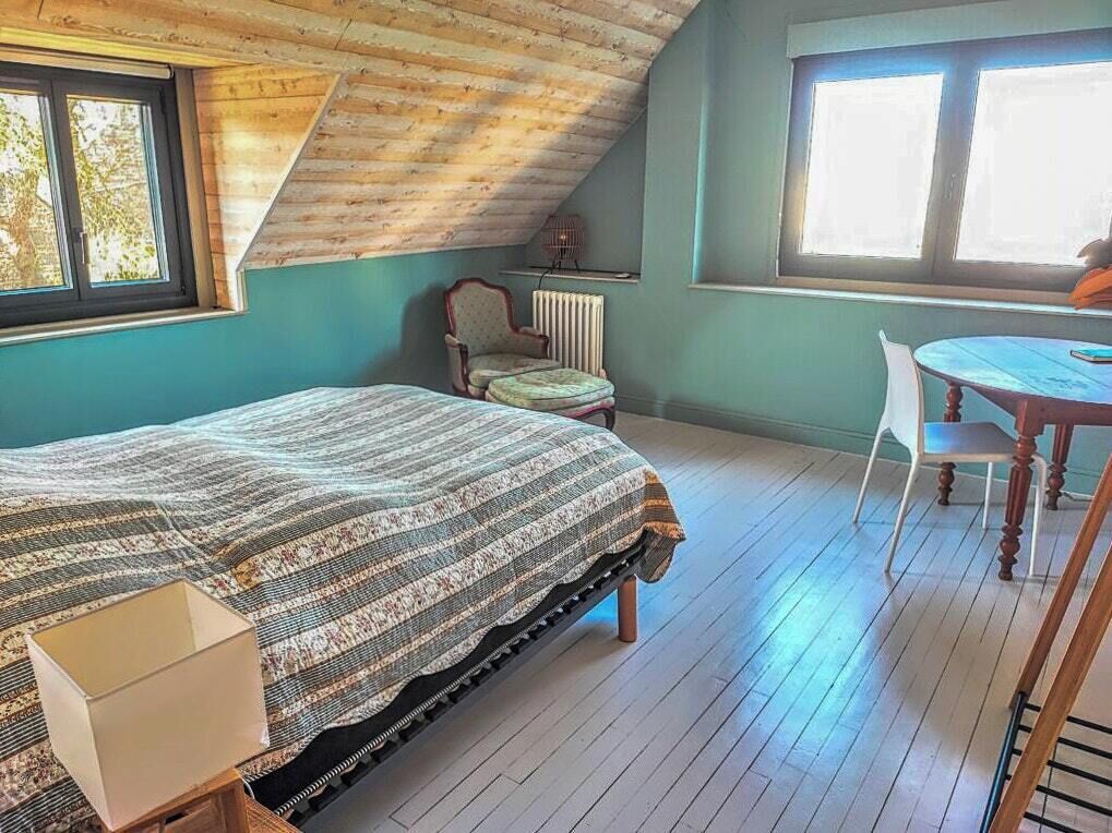 A room with a sloped ceiling, a bed with a patterned blanket, a chair, a table, and a radiator.