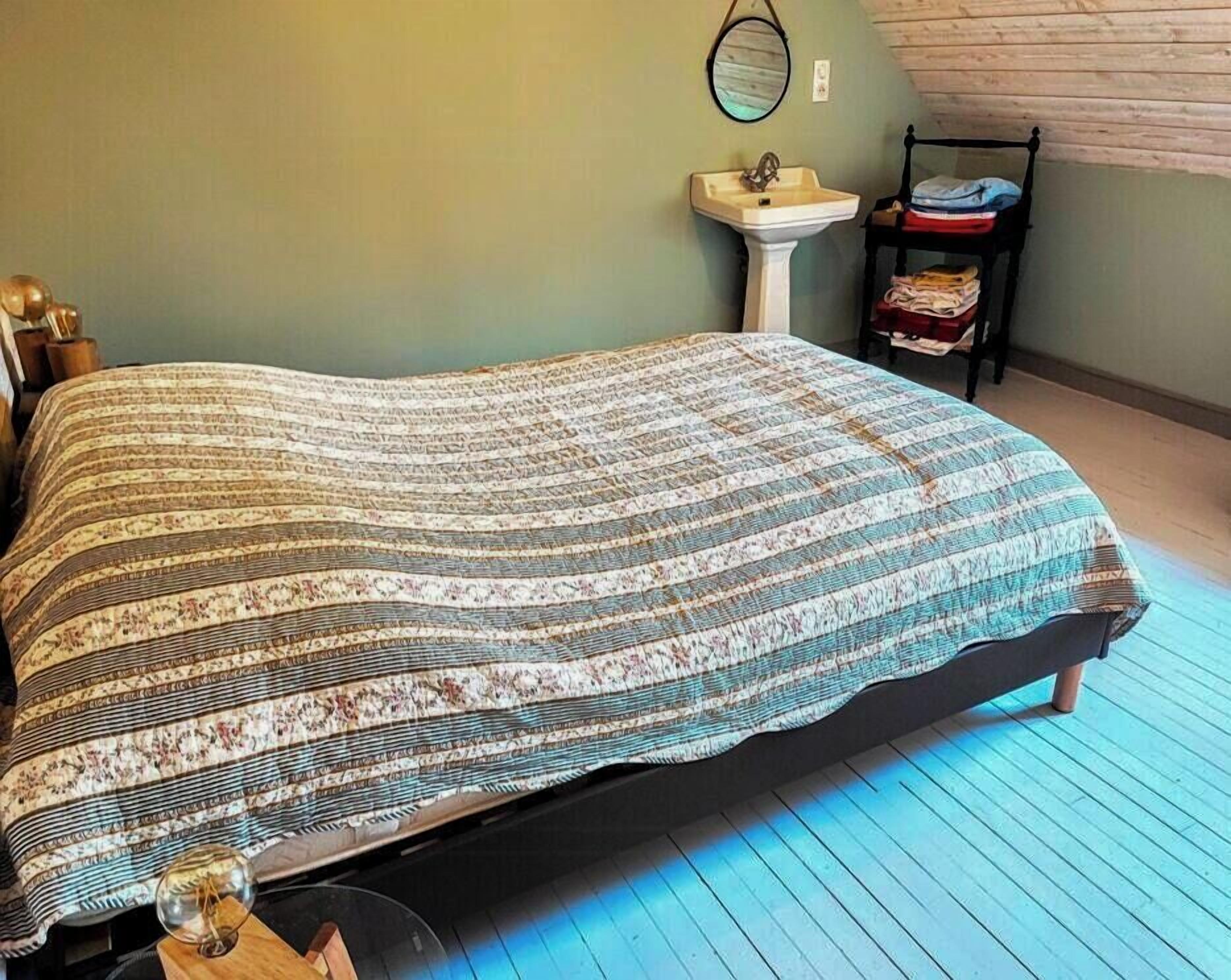 Un lit avec une couvre-lit à motifs, une table de chevet avec une lampe, un miroir fixé au mur et un petit lavabo.