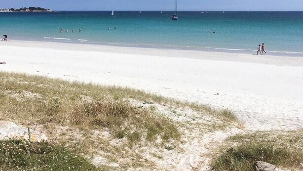 Ein Strand mit klarem türkisblauem Wasser, einem sandigen Ufer und einem entfernten Segelboot.