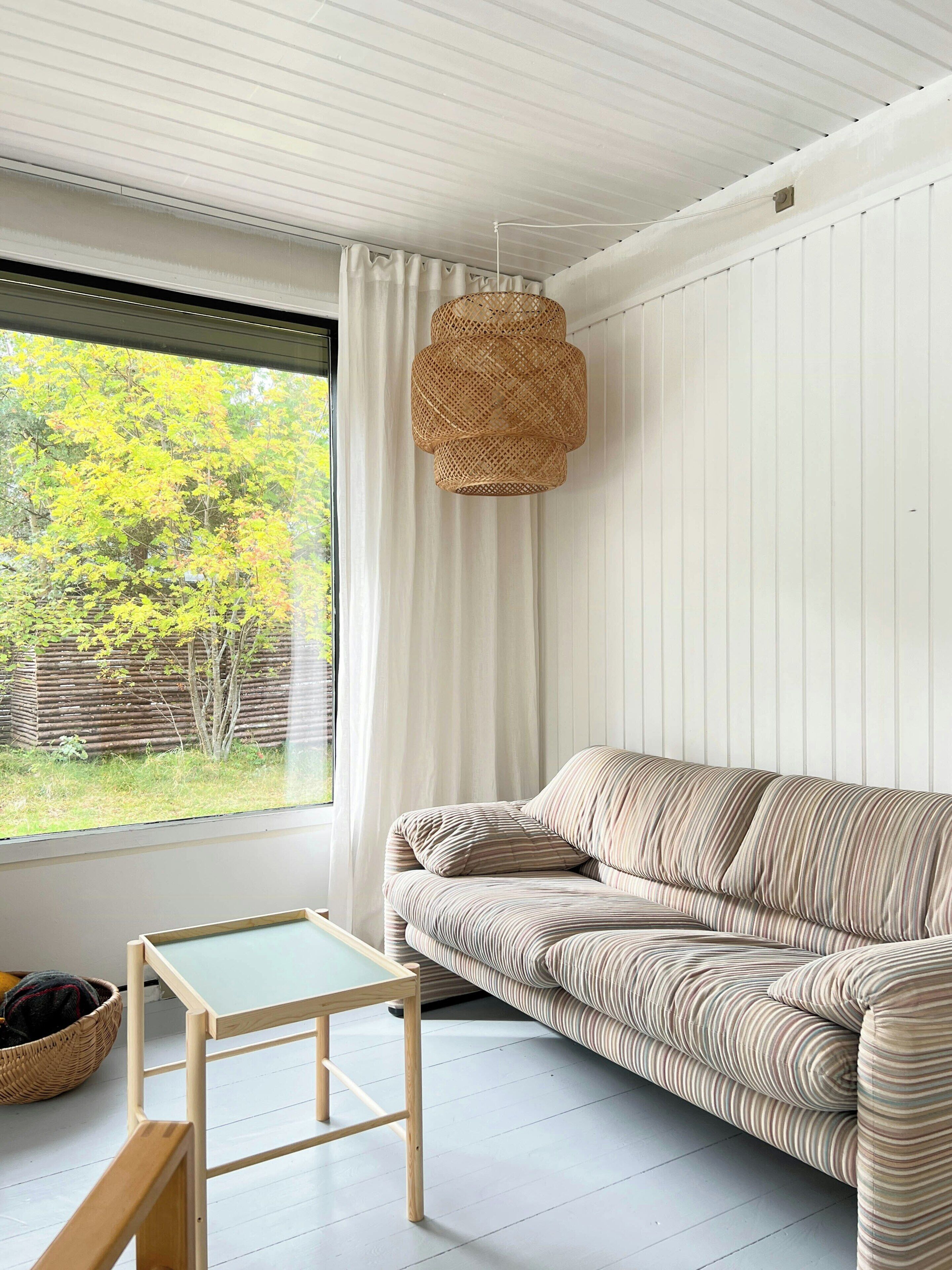 Une pièce avec un canapé, une petite table, un luminaire suspendu en osier et une vue sur les arbres à l’extérieur.