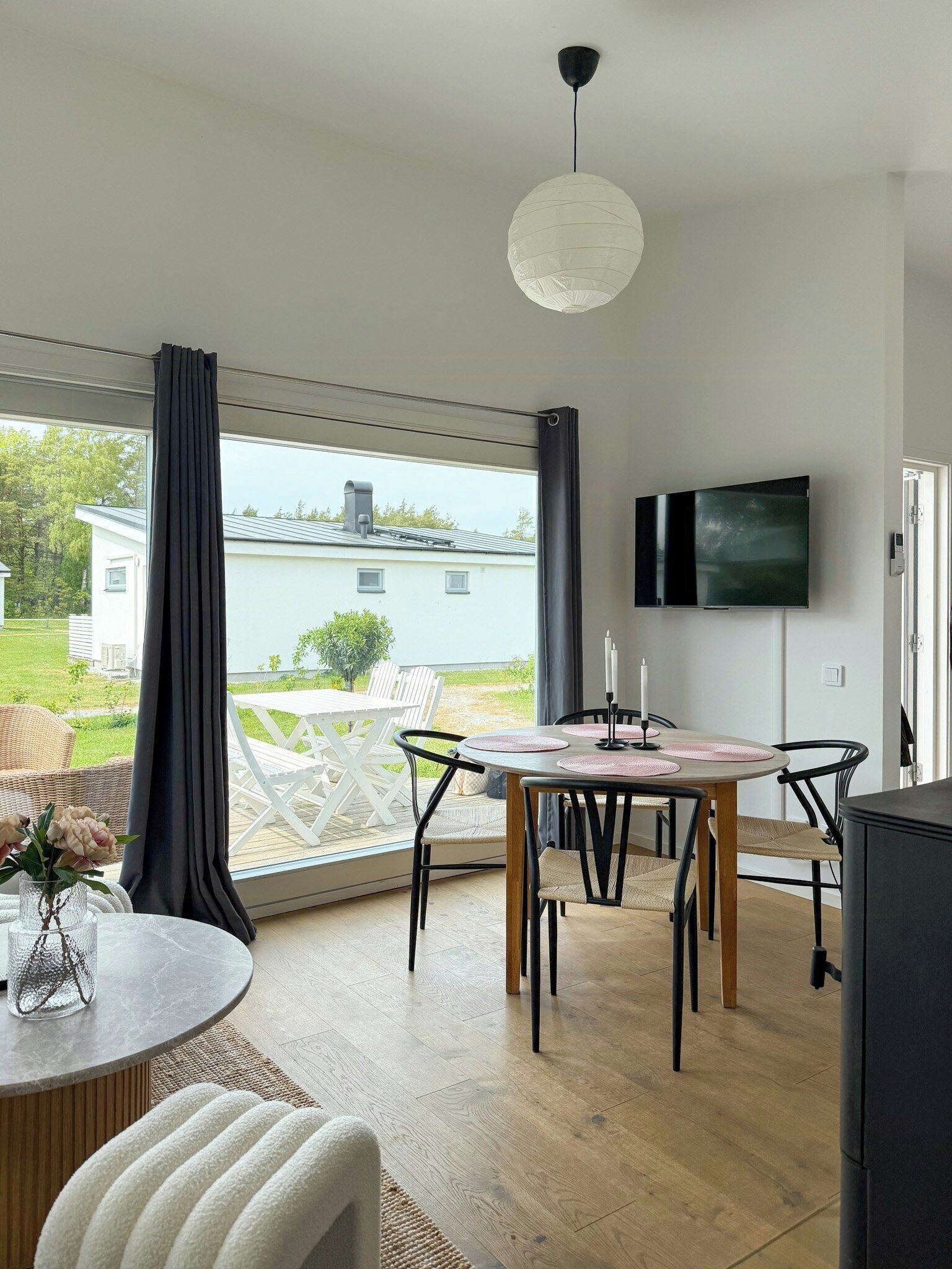 A modern dining area with a round table, chairs, a TV, and a view of a house through large windows.