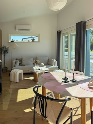 Ein modernes Wohnzimmer mit einer Couch, einem Esstisch mit rosa Platzsets, einer Lampe und Blick durch große Fenster.