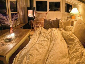 Ein gemütlicher Raum mit einer Couch, einem Tisch mit Kerzen, einem Korbsessel und einem Kaminofen.