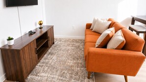 A modern living room with a flat-screen TV mounted on the wall, a wooden entertainment unit, an orange sofa with cushions, and a patterned rug.