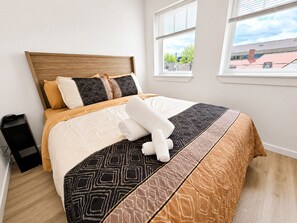 A neatly made bed with a patterned bedspread, a wooden headboard, and two windows with blinds.