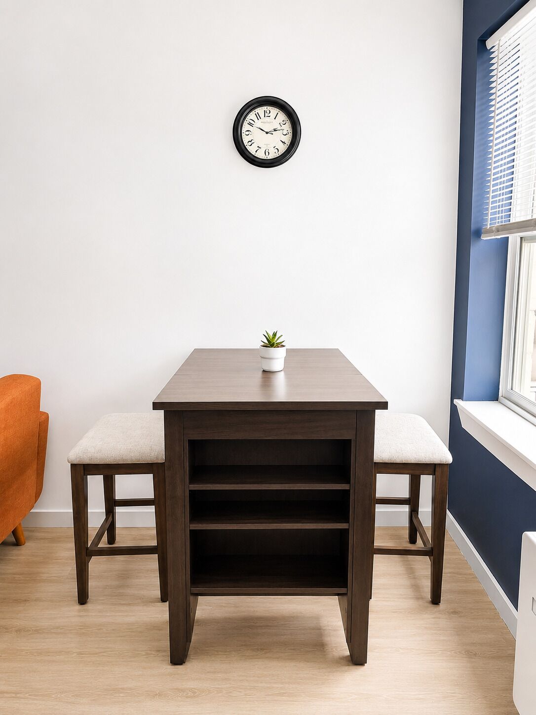 Una habitación con una mesa de madera, dos sillas, una planta en maceta, un reloj de pared y un ventanal con persianas.